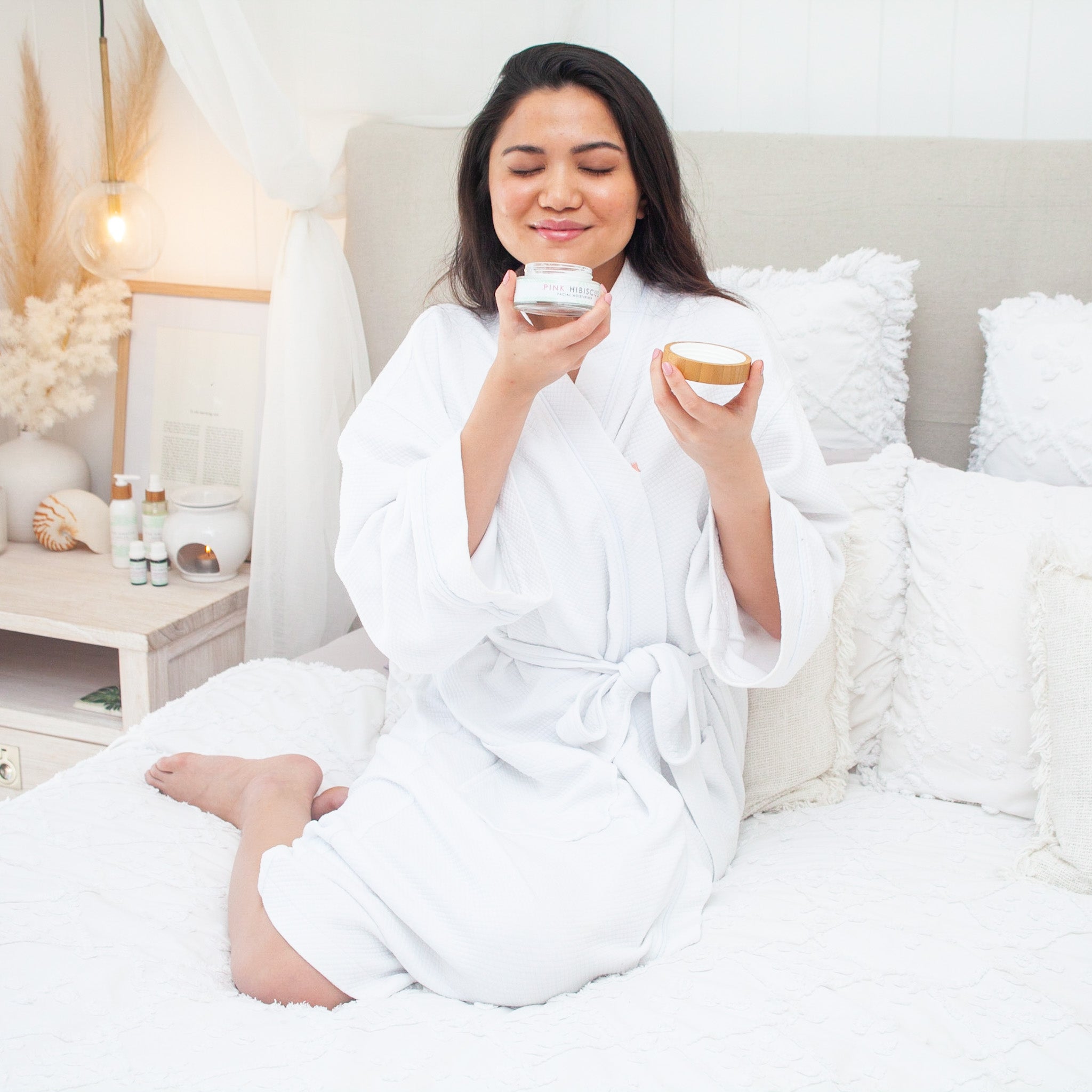 Filipino woman smelling Pink Hibiscus Facial Moisturiser in Hamptons-style bed wearing a white robe