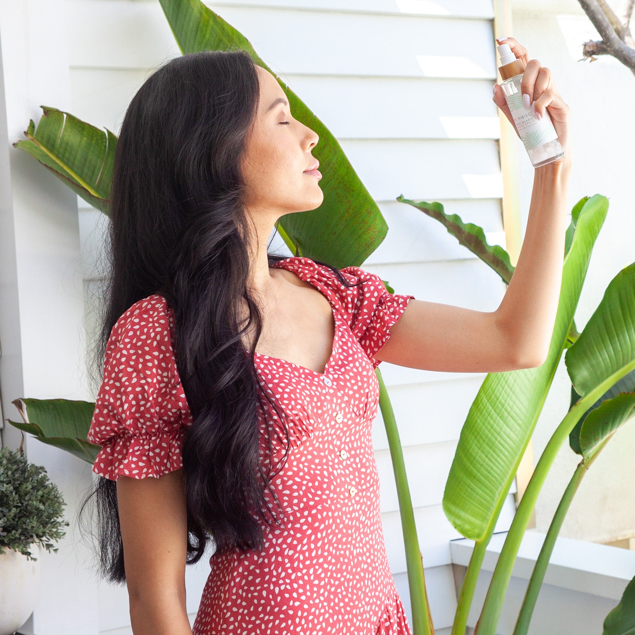 Thai woman spraying Pink Hibiscus Rose Quartz Facial Spritz onto her face in front of traveller palm